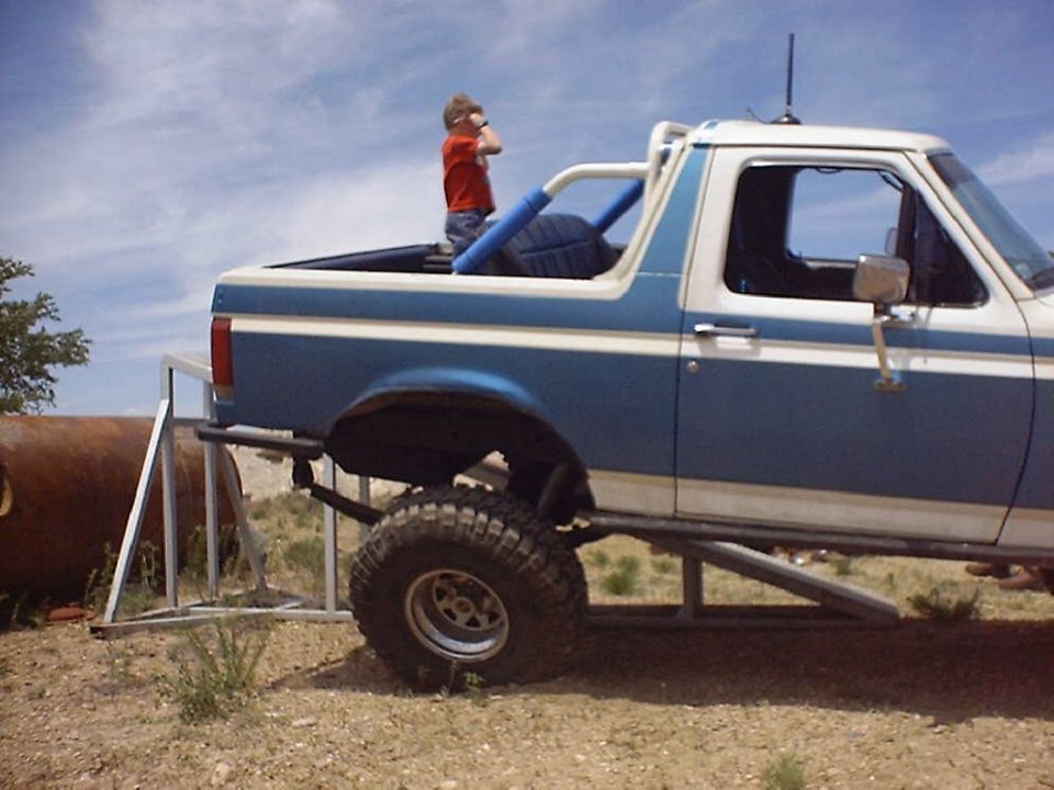 Blue and white Ford Bronco with a kid in the bed, lifted with big tires