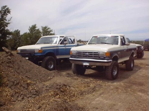 1988 Ford Bronco - Two Ford trucks are parked on a dirt lot under a cloudy sky.