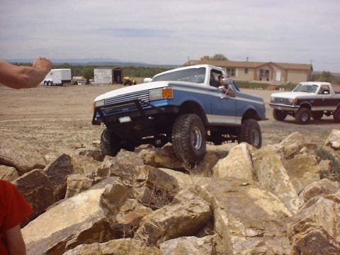 1988 Ford Bronco - Two trucks navigate a rocky terrain under a cloudy sky, with buildings in the background.