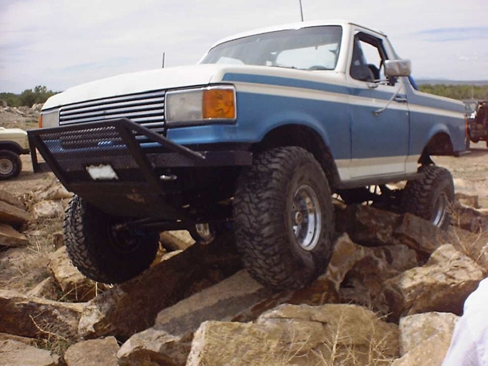 Blue and white Ford Bronco with aftermarket bumper and large tires climbing over rocks
