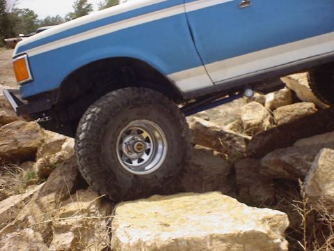 1988 Ford Bronco - A blue and white Ford Bronco navigates a rocky terrain, showing its front wheel and side profile.