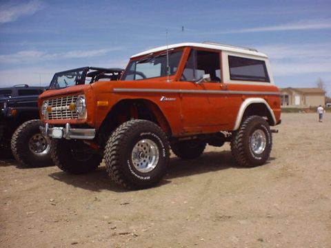 1988 Ford Bronco - A lifted orange Ford Bronco with a white hardtop sits parked on a dirt lot under a blue sky.