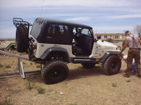 1988 Ford Bronco - A man works on the front wheel of a modified Jeep Wrangler parked on a metal ramp outdoors.
