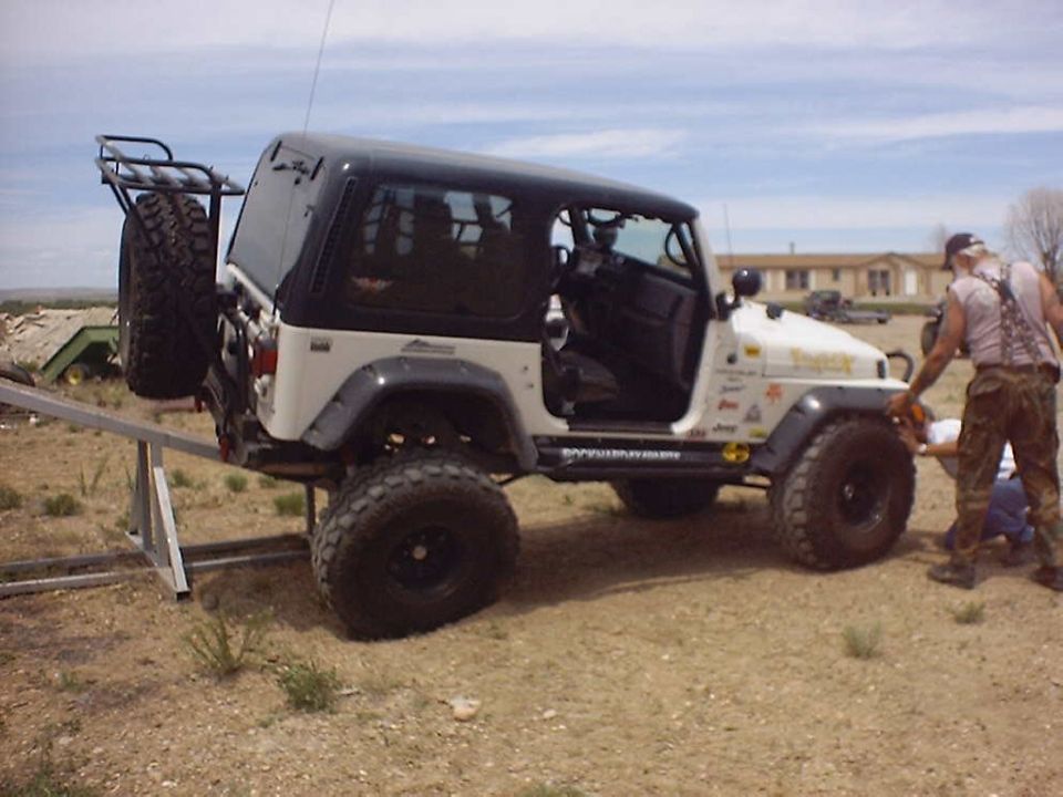 White Jeep Wrangler with black top, spare tire on rear rack, and a man working on the front wheel