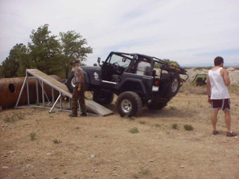 1988 Ford Bronco - A man stands by a dark blue Jeep Wrangler positioned on a ramp in an outdoor, dusty environment.