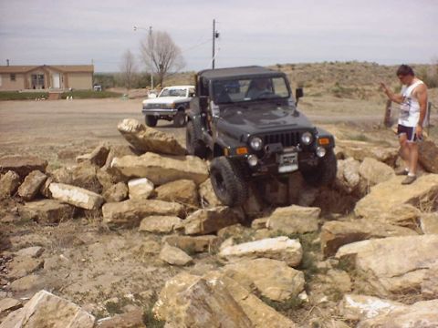 1988 Ford Bronco - Jeep Wrangler tackling an obstacle course made of rocks, with another vehicle visible behind it.