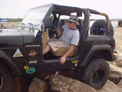 1988 Ford Bronco - A man wearing a hat and sunglasses sits in the driver's seat of a Jeep Wrangler, which is positioned on a rocky terrain.
