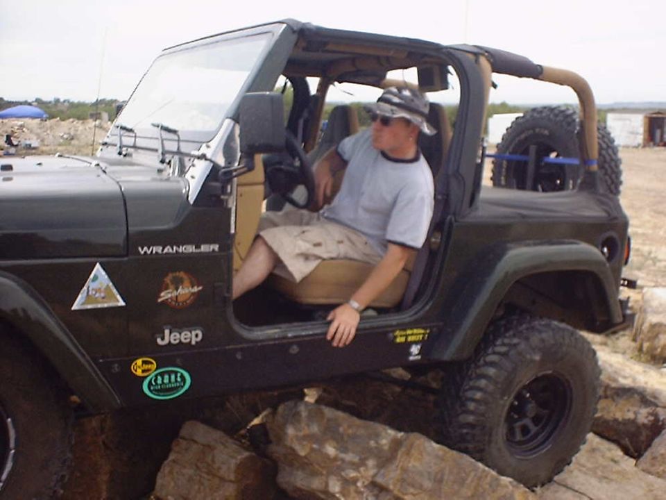 Man in hat sits in a dark green Jeep Wrangler parked on rocks