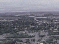 1998 Mercury Sable - A wide aerial view shows a town inundated by floodwaters from a nearby river under an overcast sky.