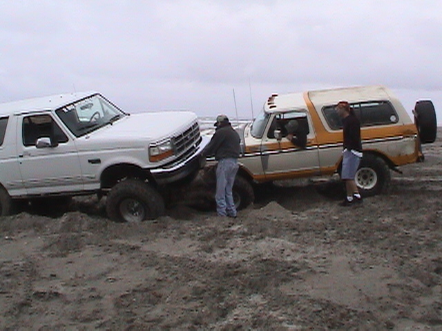 White Ford truck and orange Bronco stuck in wet beach sand with two men working