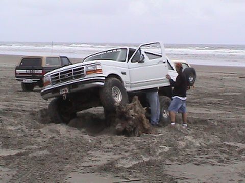 1994 Ford Explorer - A white Ford Bronco is stuck in the sand on a beach, with two people attending to it.