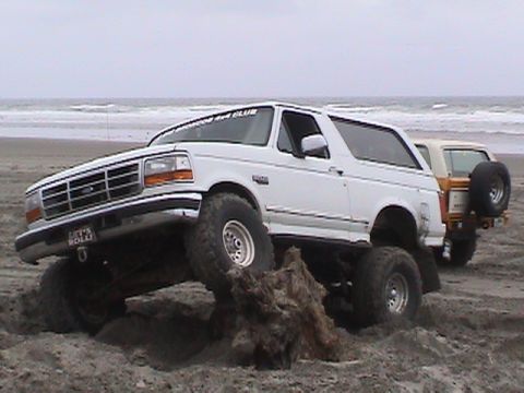 1994 Ford Explorer - A white Ford Bronco navigates sandy terrain on a beach, with another vehicle visible in the background.