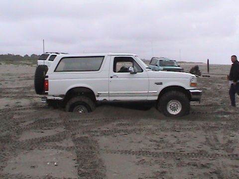 1994 Ford Explorer - A white Ford Bronco is stuck in the sand on a beach, with another vehicle visible in the background.