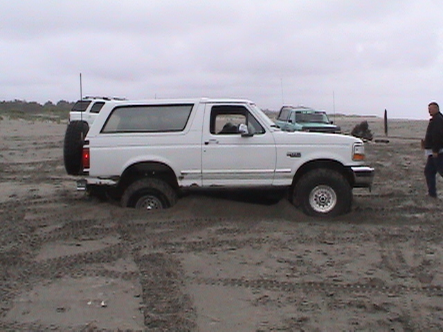 White Ford Bronco with rear-mounted spare tire parked on a tire-tracked beach