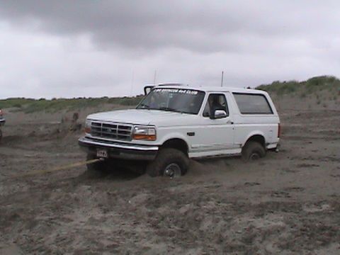 1994 Ford Explorer - A white Ford Bronco is stuck in the sand with a tow strap attached, while another vehicle is partially visible in the background.
