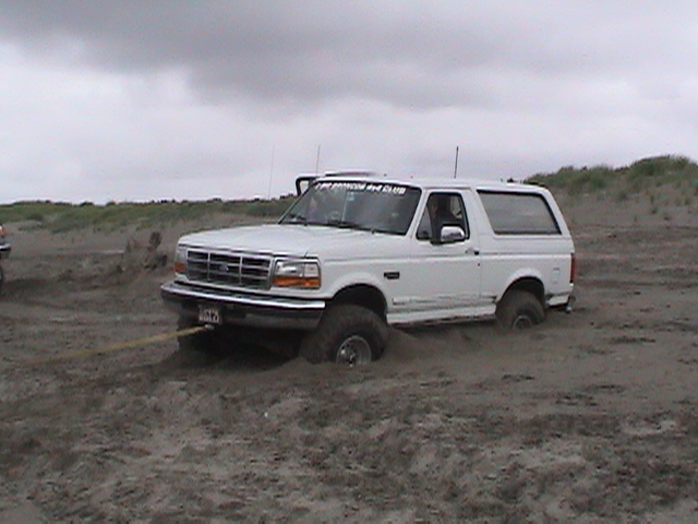 White Ford Bronco stuck in sand, front passenger side buried. Tow strap attached to front bumper. "PORT ANGELES 4X4 CLUB" sticker on windshield