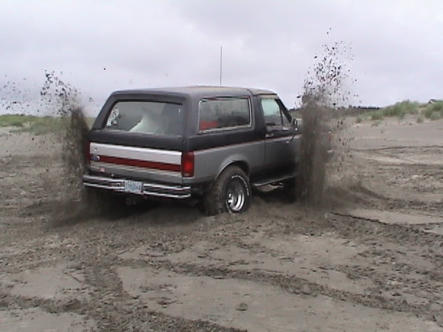 Gray Ford Bronco spraying sand and mud from its tires in a sand pit, rear view