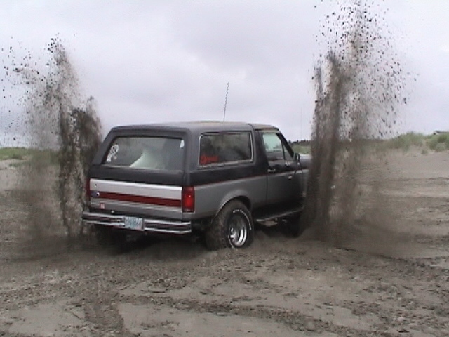 Gray Ford Bronco spraying twin rooster tails of sand from its rear tires on a beach