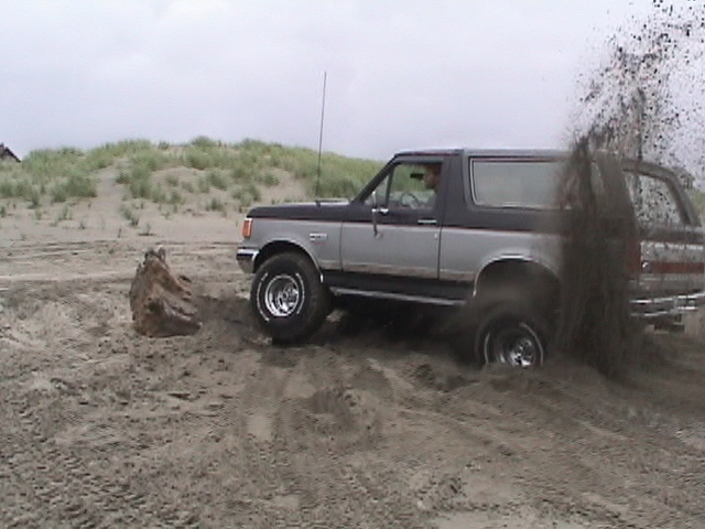 Two-tone gray Ford Bronco spinning tires and throwing sand on a beach dune
