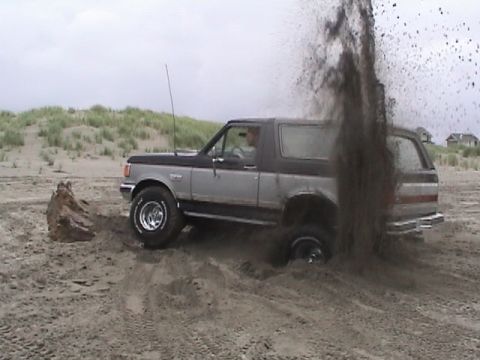 1994 Ford Explorer - A two-tone Ford Bronco is stuck in the sand, with a plume of dirt erupting from its rear wheel.