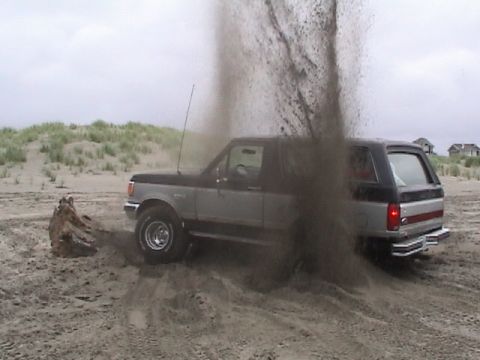 1994 Ford Explorer - Ford Bronco is stuck in the sand on a beach, with a large plume of dust rising.