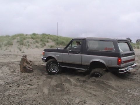 1994 Ford Explorer - A two-tone Ford Bronco is partially submerged in sand near a dune.