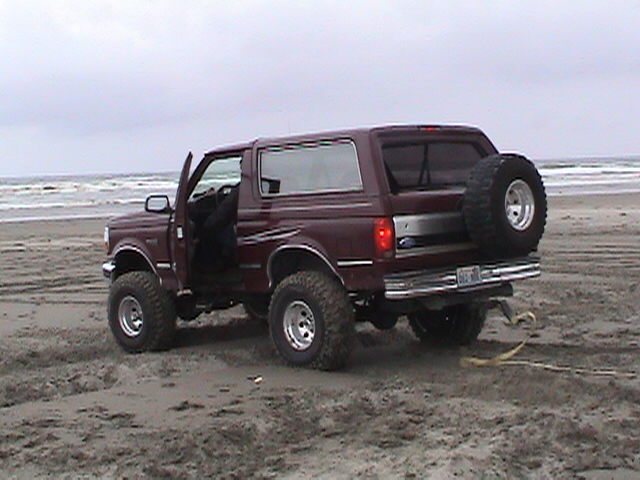 Lifted maroon two-door Ford Bronco with door open on a beach, rear three-quarter view