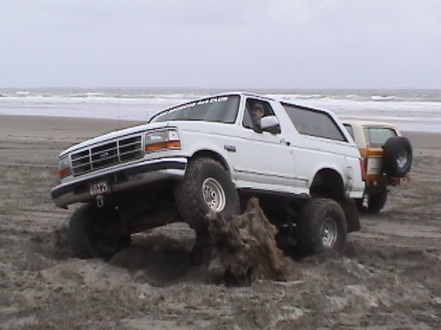White lifted Ford Bronco climbing a driftwood stump on an ocean beach with surf behind