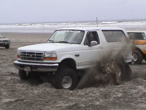 1994 Ford Explorer - A white Ford Bronco is stuck in the sand on a beach, with another vehicle partially visible behind it.