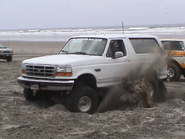 White Ford Bronco spinning tires in wet beach sand, ocean behind
