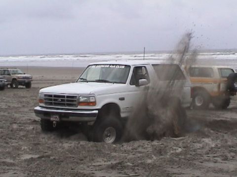 1994 Ford Explorer - A white Ford Bronco is stuck in the sand on a beach, kicking up dirt.
