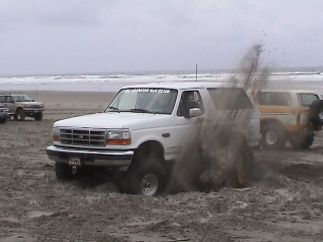 White Ford Bronco spinning tires and throwing sand on an ocean beach, 3/4 front view