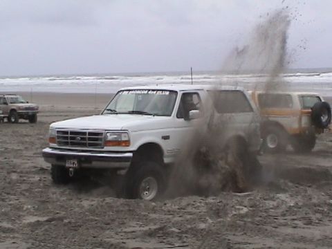 1994 Ford Explorer - A white 1994 Ford Bronco is stuck in the sand on a beach, with another Bronco visible in the background.
