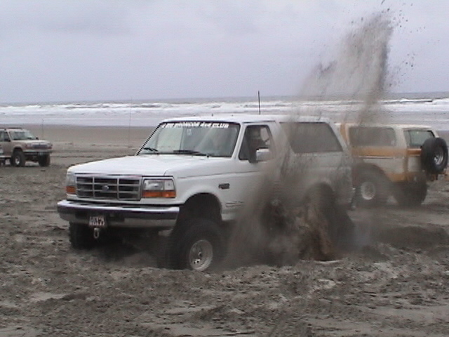 White Ford Bronco spraying sand through a beach mud hole with surf and other 4x4s behind