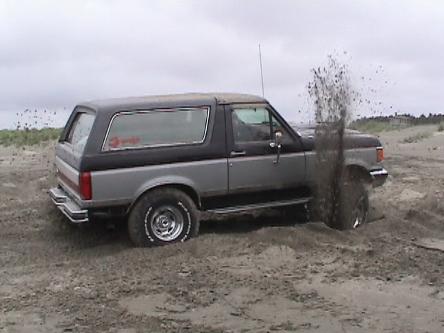 Gray and black Bronco spinning its tires and spraying sand on an overcast beach