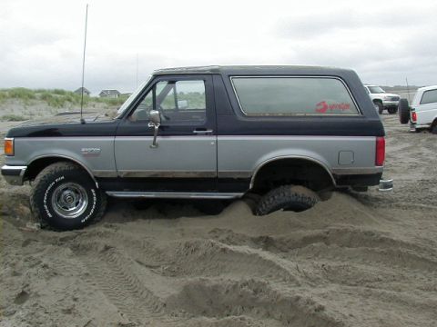 1994 Ford Explorer - A two-tone Ford Bronco is stuck in the sand with its rear tire partially buried.