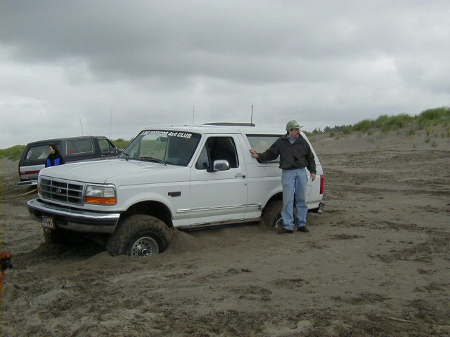 White Ford Bronco stuck axle-deep in beach sand with man at open driver's door