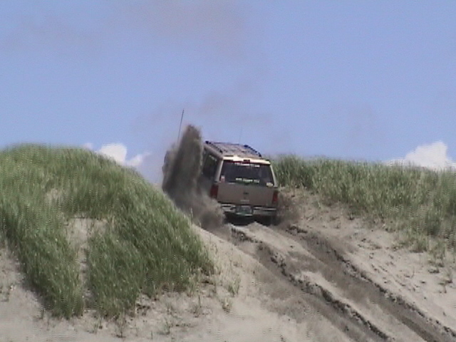 Sand-covered SUV climbing a dune, throwing sand, roof light bar and whip antenna