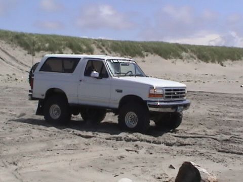 1994 Ford Explorer - A white Ford Bronco is driven on a sandy beach, with dunes and a cloudy sky overhead.