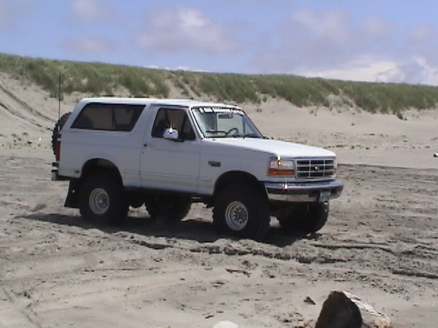 Lifted white full-size Ford Bronco parked on sand beside beach grass dunes
