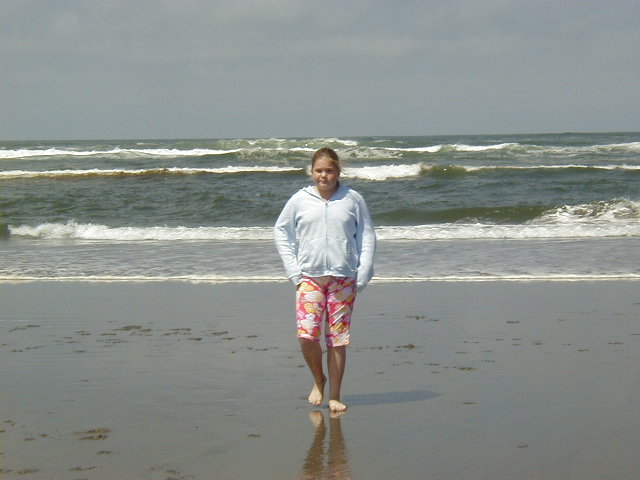 Person in gray hoodie and floral shorts walking barefoot on a beach with waves behind