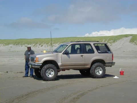 1994 Ford Explorer - A man stands beside a lifted 1994 Ford Explorer on a sandy beach under a partly cloudy sky.