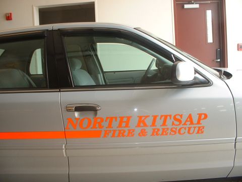 1999 Ford Crown Victoria - Side view of a silver sedan with "NORTH KITSAP FIRE & RESCUE" text and an orange stripe.