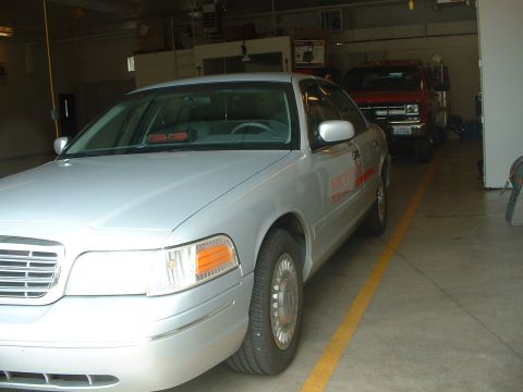 1999 Ford Crown Victoria - A silver sedan is parked in a garage, with a red truck visible behind it.