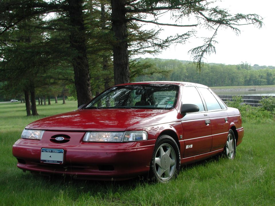 Red Ford Taurus SHO sedan parked on grass among pine trees by water