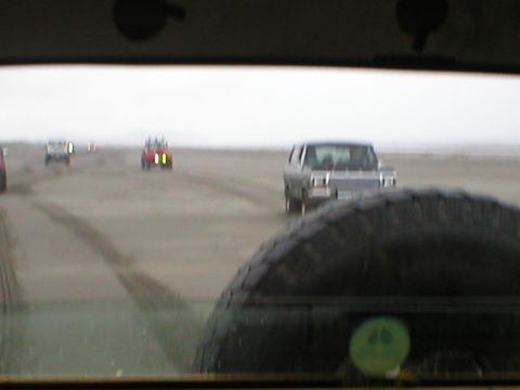1978 Ford Bronco - Several vehicles drive across a wide, sandy beach under an overcast sky.