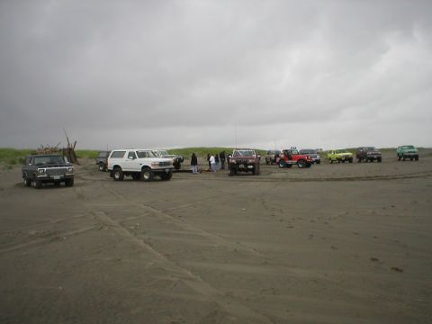 1978 Ford Bronco - A group of off-road vehicles gathered on a sandy beach, with people standing near some of them.