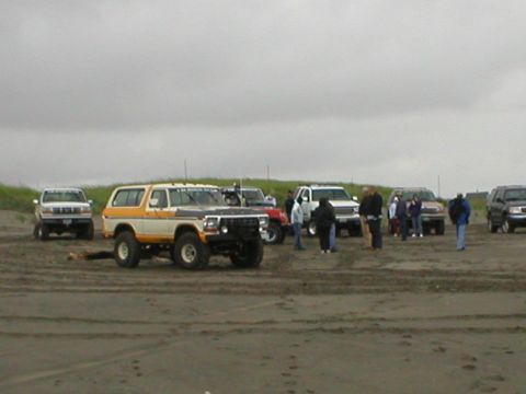 1978 Ford Bronco - A group of people and several off-road vehicles gather on a sandy beach under an overcast sky.
