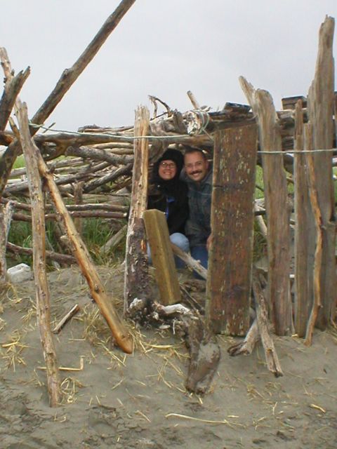 1978 Ford Bronco - Couple poses inside a driftwood shelter on a beach, with a cloudy sky overhead.