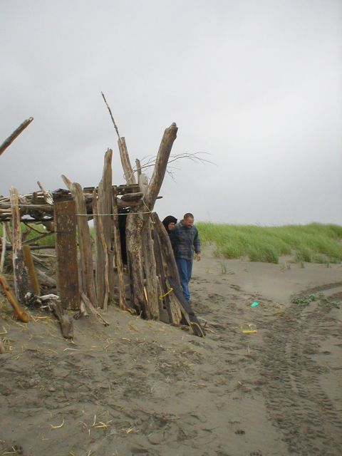 1978 Ford Bronco - Two people stand near a crude shelter made of driftwood on a cloudy beach.